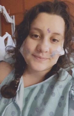 A young adult takes a selfie in the hospital while wearing a hospital gown. Thin tubing is taped to their face and goes up their nose. They have a purple mark on their forehead, between their eyebrows, indicating where their pituitary tumor is.