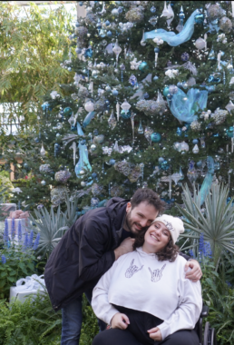 A photo of a young couple smiling outside in front of a large Christmas tree. Noura is sitting in a wheelchair wearing a pink sweatshirt and beanie, while their partner, Peter, is standing behind them and hugging their shoulders.