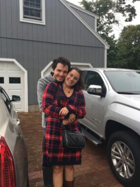 Noura and Peter Costany stand outside in a driveway between two cars hugging. Peter is wearing a gray sweatshirt and Noura is wearing a plaid knee-length dress.