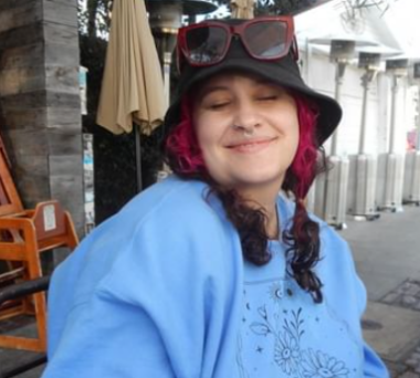 A person in their late 20s smiles with their eyes closed for a photo while seated outside at what appears to be a restaurant. They're wearing a black hat with sunglasses balanced on top of it, and a blue sweatshirt. Their hair is pulled into low pigtails, and while most of it appears to be dark brown, the money pieces are dyed hot pink. 