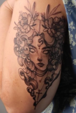 A close-up photo shows a black-and-white tattoo on the back of a person's upper arm. It depicts a person with curly hair and white eyes surrounded by flowers.
