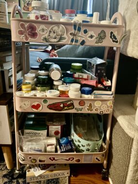 A pink, three-shelf utility cart with wheels is full of medications and other medical gear and has decorative stickers all over it.