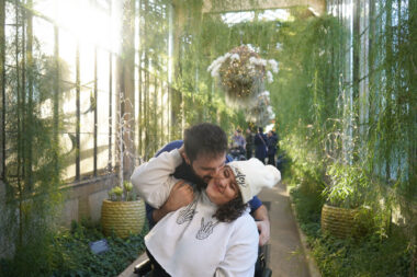 A couple pauses for a photo in what appears to be a greenhouse. The husband is leaning down and kissing his partner, who is in a wheelchair, on the cheek.